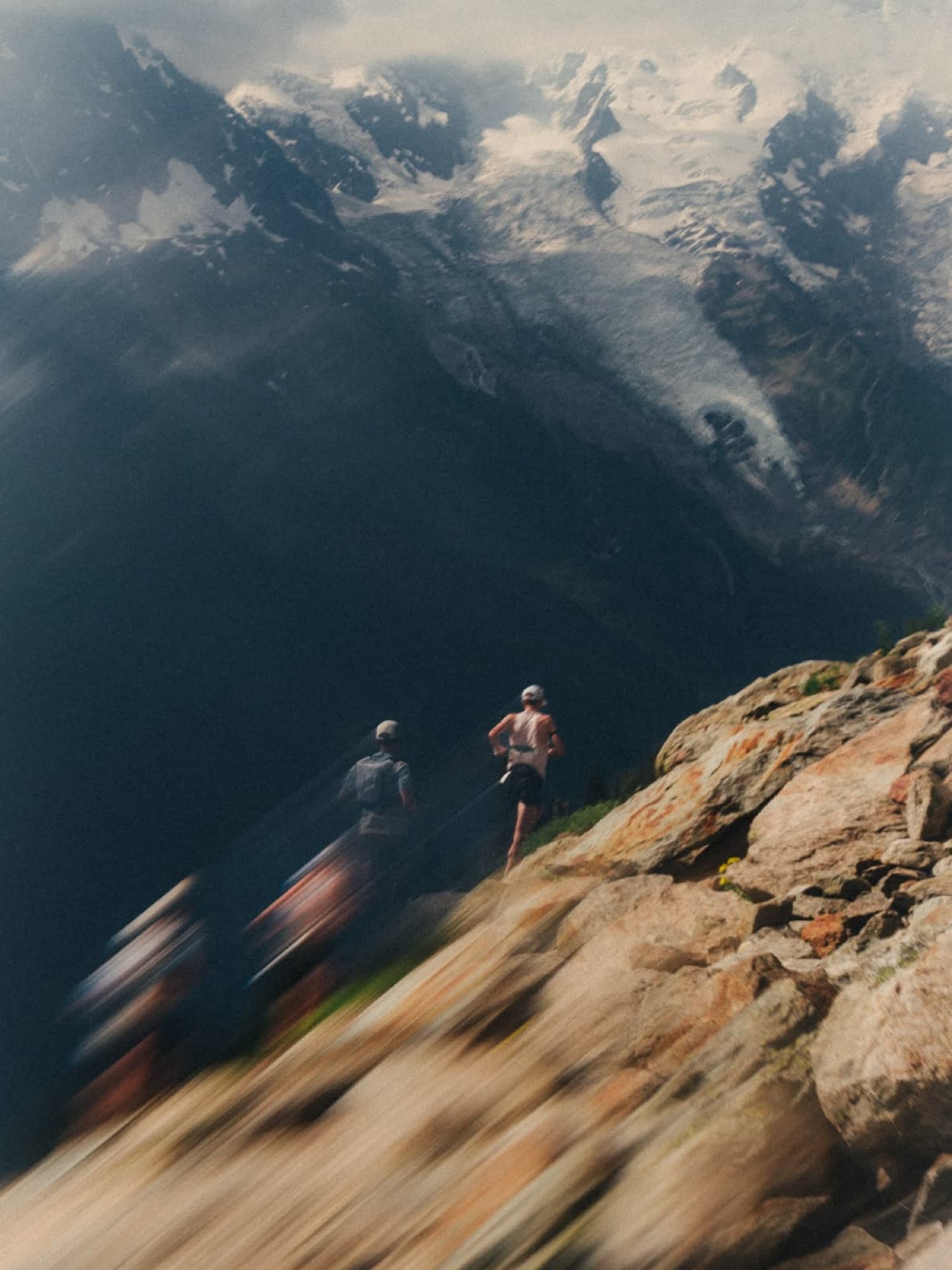 Two trail runners moving along a rocky ridge beneath glaciated peaks